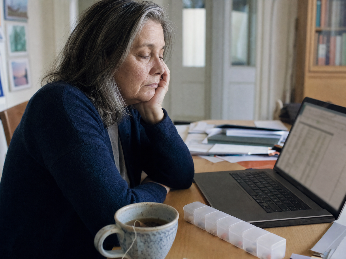 A woman at a desk in late afternoon light, chin in hand, a pill box beside the keyboard